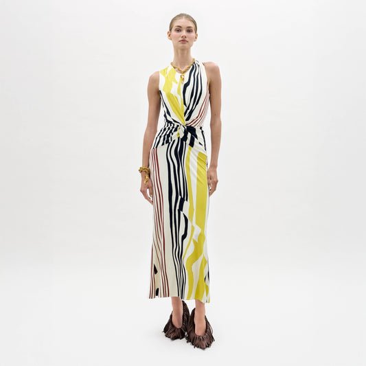 A woman poses against a white background, wearing the Luna Dress—an Italian fabric midi with abstract yellow, black, and white stripes, side waist cutout, criss-cross back straps—and brown fringed heels. She completes the look with gold jewelry.
