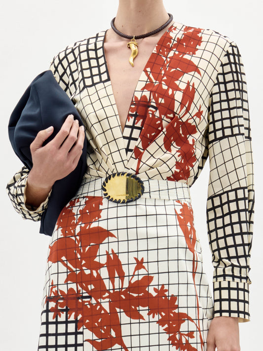 A woman stands against a white background wearing the Kadira Dress, a checked dress with red leaf prints, paired with a black clutch and brown fringed shoes.