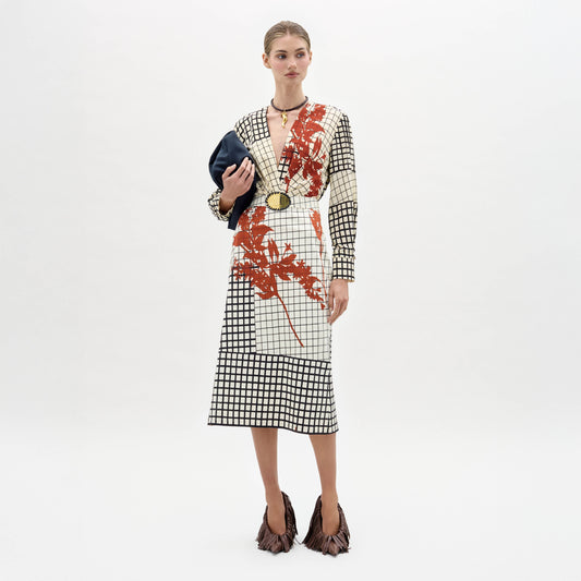 A woman stands against a white background wearing the Kadira Dress—an A-line, knee-length, long-sleeve style with a black and white grid pattern and red-orange botanical print—paired with fringed brown shoes and holding a black clutch.
