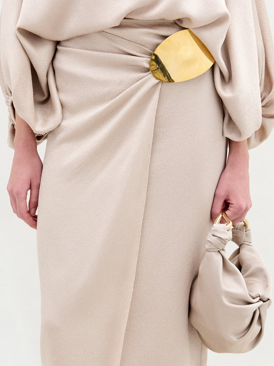A woman stands against a white background wearing the beige Adrianne Skirt with an off-the-shoulder hammered satin top, a gold-tone embellishment belt, gold heels, and carrying a matching beige handbag.