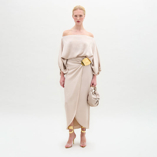 A woman stands against a white background wearing the beige Adrianne Skirt with an off-the-shoulder hammered satin top, a gold-tone embellishment belt, gold heels, and carrying a matching beige handbag.