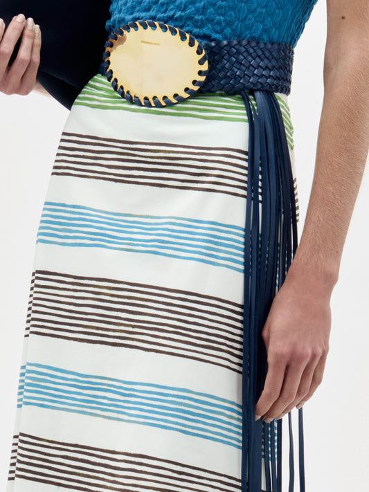 A person wears the Lyla Skirt, featuring white fabric with blue, green, and brown horizontal stripes, paired with a blue textured top and a wide dark blue woven belt with a large oval buckle. Their right hand rests at their side.