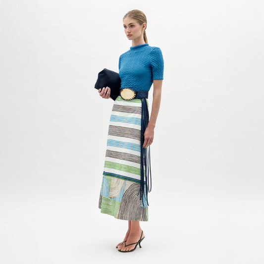 A woman stands against a white background wearing a blue short-sleeve top, the Lyla Skirt—featuring a fitted striped midi design in blue, green, and brown—a wide black belt with gold buckle, black sandals, and holding a black clutch.