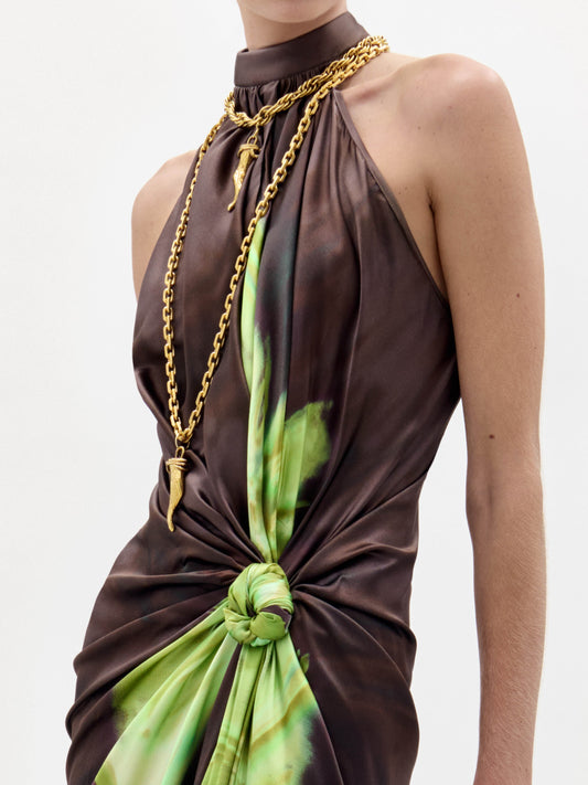 A woman in the Tizziana Dress, a brown and green sleeveless style, holds a large fringed bag and wears black heeled sandals against a plain white background.