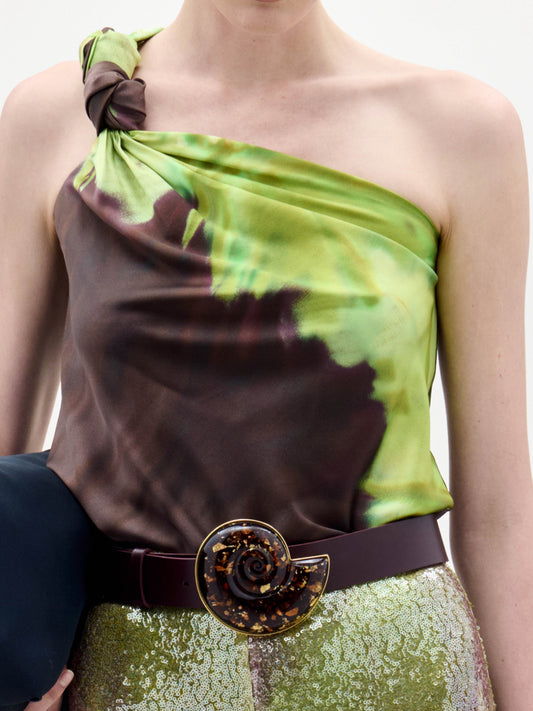 A person in the Yanet Blouse, a green and brown tie-dye silk top with an asymmetric neckline and side knot, paired with gold textured pants and a wide belt with a standout spiral buckle.