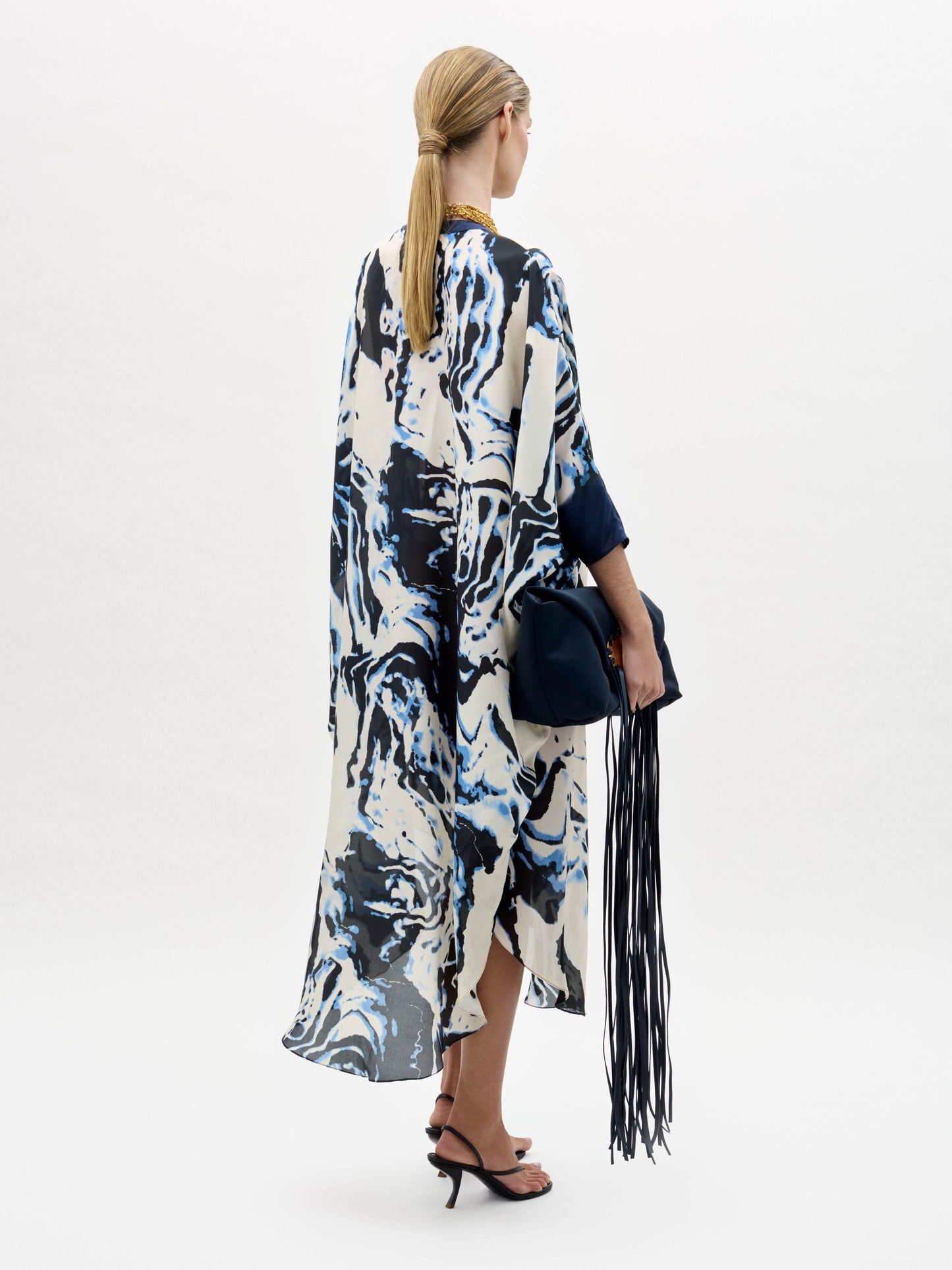 A woman stands against a plain background wearing the Elea Tunic with an abstract pattern, black sandals, gold jewelry, and holding a navy fringe bag.