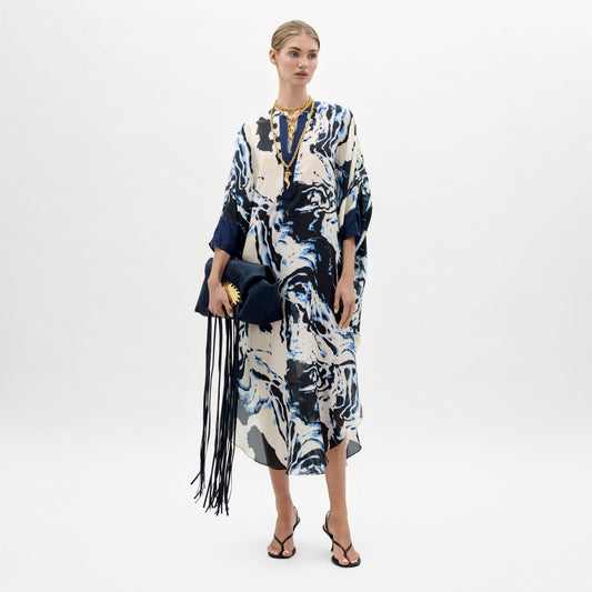 A woman stands against a plain background wearing the Elea Tunic with an abstract pattern, black sandals, gold jewelry, and holding a navy fringe bag.