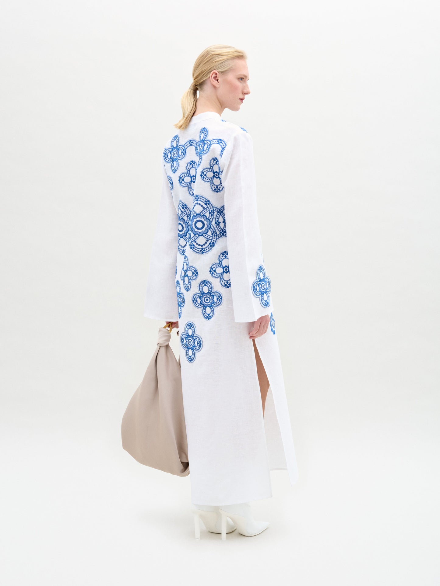 A woman stands against a white background wearing the Adalys Tunic, an Italian-fabric maxi with blue circular patterns and flared bell sleeves, styled with white boots and a large beige bag.