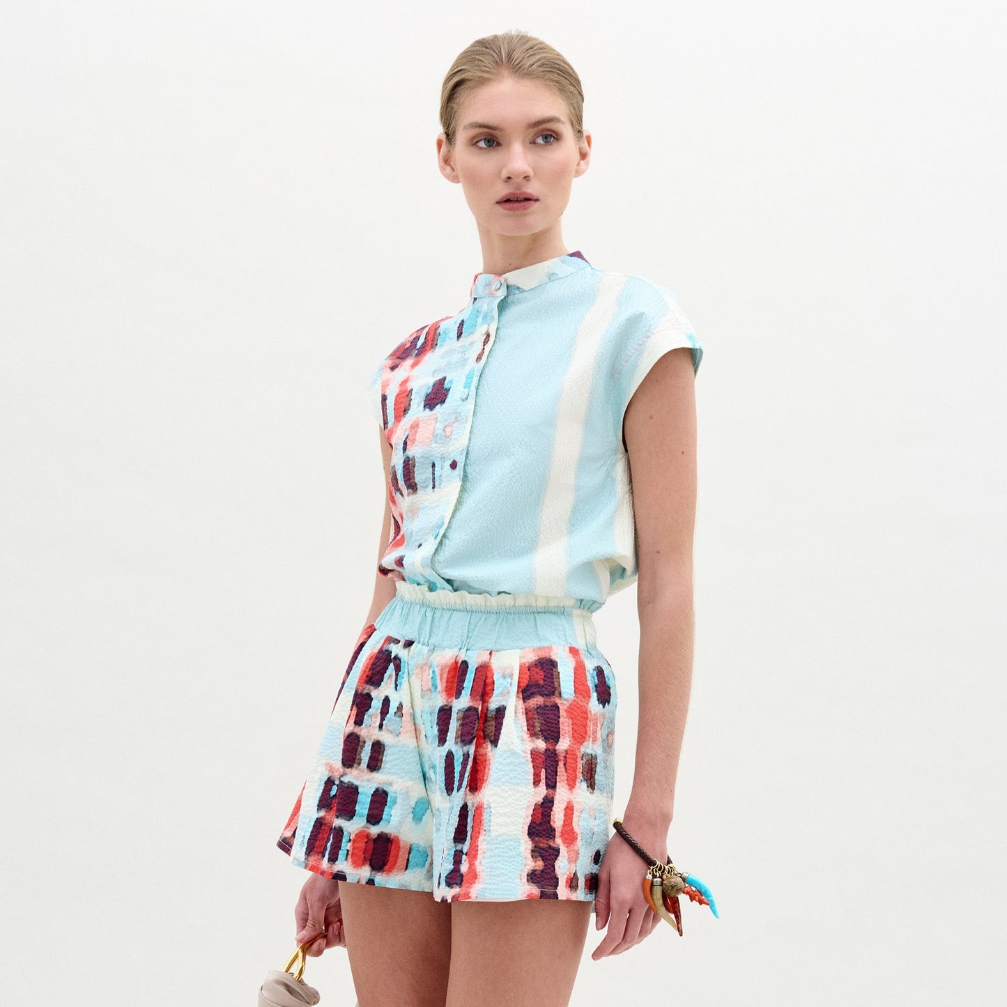 Wearing the Adele Blouse, a woman pairs its light blue and multicolored patterned collarless design with matching shorts made from Italian fabric, holding a small bag as she stands against a plain light background.