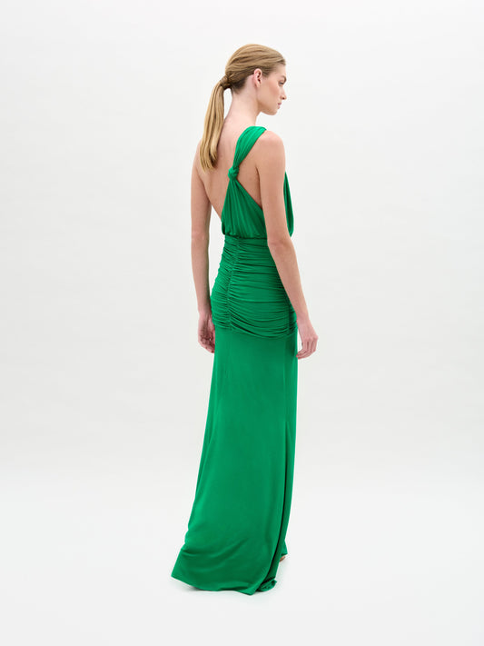 A woman stands against a plain white background wearing the Antara Dress—a bright green, fitted maxi with an asymmetric one-shoulder neckline, gathered waist, and front slit.
