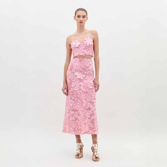 A woman stands wearing the Alida Dress Set Pink, a chic two-piece floral lace outfit featuring a crop top and a mid-length skirt, paired with high-heeled shoes adorned with decorative elements. Perfect for an elegant event, this set is guaranteed to impress; make sure to order by the ship-by date of February 1st, 2025.