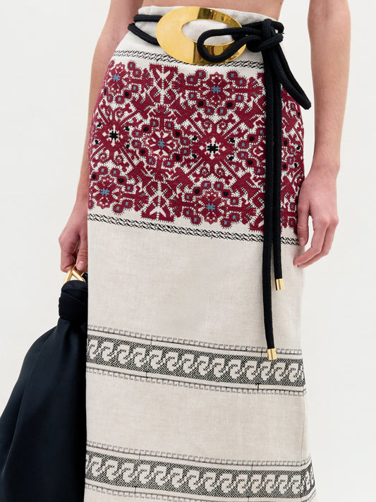 A woman stands against a plain background wearing the Atira Skirt, styled with a black belt, gold sandals, and carrying a large black bag.