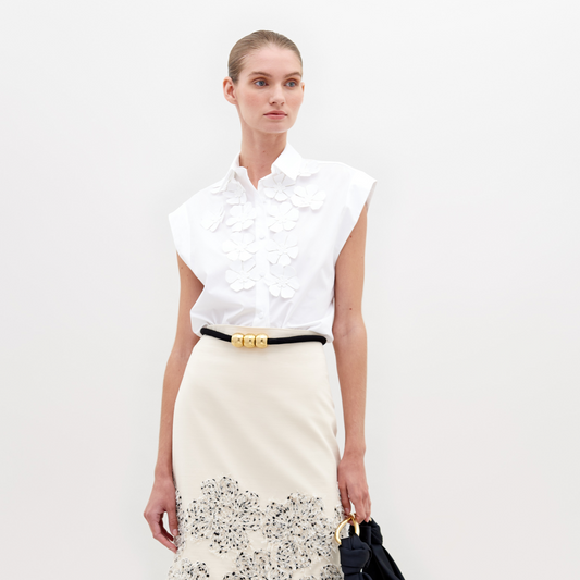 The individual is dressed in an Azalea Blouse, a white sleeveless top with floral appliqué, paired with a cream skirt featuring a patterned hem, and holds a black bag against a plain background.