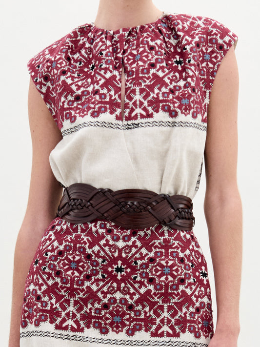 A woman stands against a white background wearing the Badra Dress, styled with a woven leather belt, beige strappy sandals, and holding a large beige bag.