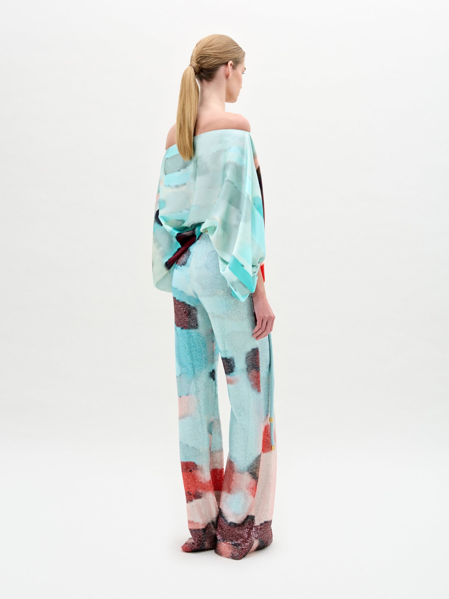 A woman stands against a plain light background wearing the Dalal Pant—high-waisted, wide-leg pants in luxurious Italian fabric with an abstract pattern—styled with a colorful off-the-shoulder top and finished with a yellow belt accent.