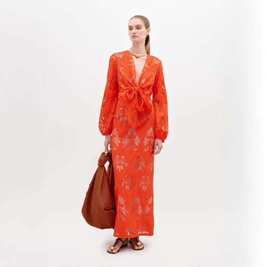 A person stands in a long, bright orange Battia Dress Rouge Embroidery with floral patterns and a front bow tie. They carry a large, rust-colored bag and wear gold jewelry and black sandals. To ensure you receive this stunning ensemble by the desired ship-by date, consider placing your pre-order for 2024 today.