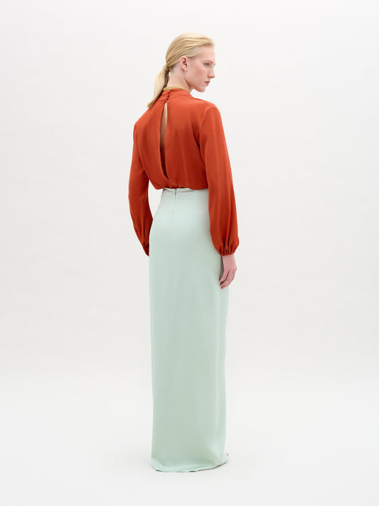 A woman wearing the long-sleeve rust-colored Delfina Blouse, a light blue skirt with a bold metallic waist accent, and layered gold necklaces stands against a plain white background.