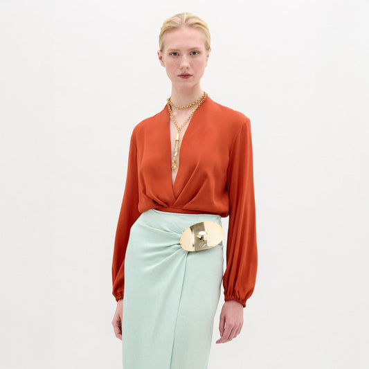 A woman wearing the long-sleeve rust-colored Delfina Blouse, a light blue skirt with a bold metallic waist accent, and layered gold necklaces stands against a plain white background.