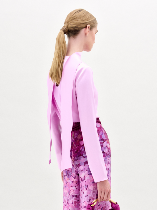 A woman poses against a plain background in the Gaby Blouse, a light pink silk long-sleeve top with a front slit, paired with high-waisted pink and purple patterned pants, a wide belt, and a small matching handbag.