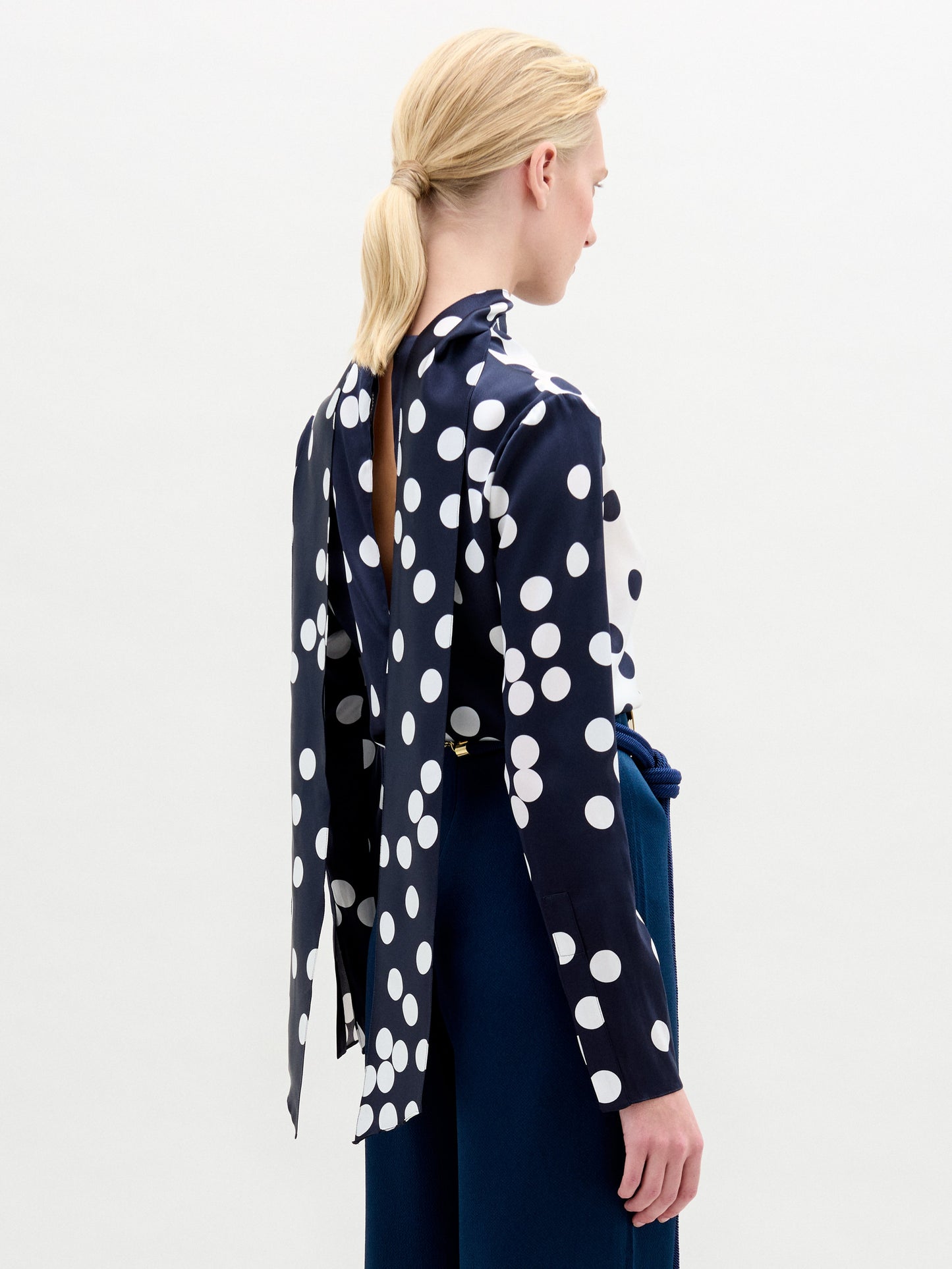 A woman stands against a plain background, wearing the Gaby Blouse—a navy and white polka dot silk top with a keyhole neckline—paired with high-waisted navy pants featuring a large oval belt buckle.