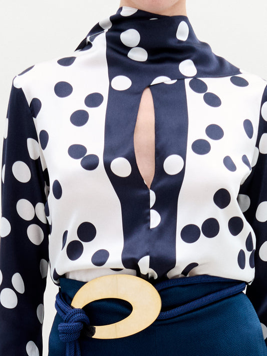 A woman stands against a plain background, wearing the Gaby Blouse—a navy and white polka dot silk top with a keyhole neckline—paired with high-waisted navy pants featuring a large oval belt buckle.
