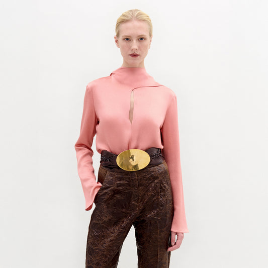 A woman stands against a plain background wearing the Gaby Blouse, a pink high-neck top with a front slit, paired with brown textured pants and a wide belt featuring a large gold oval buckle.