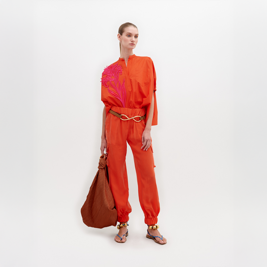 A person standing in a white space wearing an orange long-sleeve top, Jess Pant Tangerine with cargo pockets, and sandals with bead embellishments. They look downwards with a neutral expression. Pre-order now to ship by November 15th.