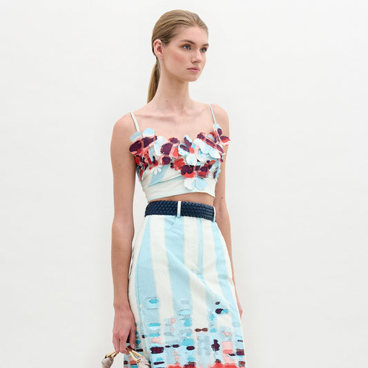 A woman stands against a plain background wearing the Kim Top— a sleeveless crop top with striking 3D floral detailing in red and blue—paired with a light blue patterned skirt.
