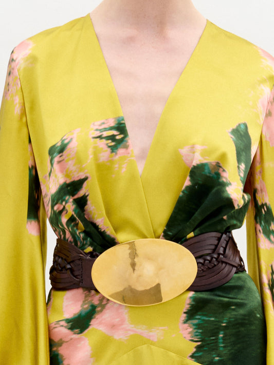 A person in a Lima Dress with a floral yellow and green pattern, a wide belt, holding a beige handbag, and wearing strappy high heels.