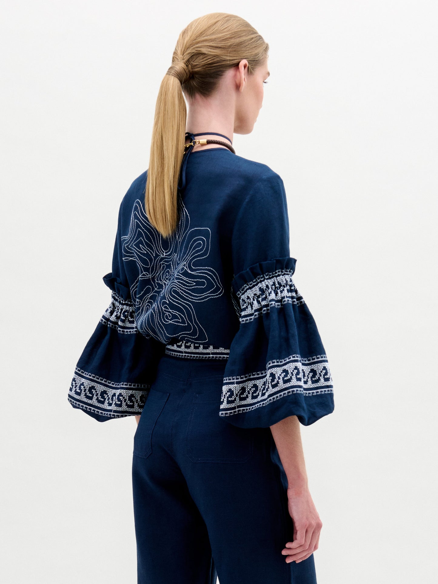 A woman stands against a plain white background wearing the Lucaya Blouse—an embroidered navy blue linen top with voluminous sleeves—paired with matching navy trousers.