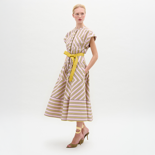 A woman models the Macarena Dress—a light pink and yellow geometric midi made from Italian fabric—styled with a woven leather belt and high-heeled sandals, set against a plain white background.