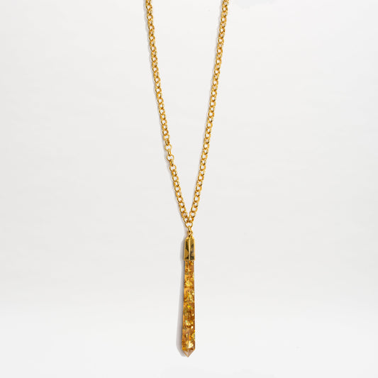 The Montse Necklace is a gold chain layering piece with a narrow geometric crystal pendant set with yellow gemstones, shown on a plain white background.