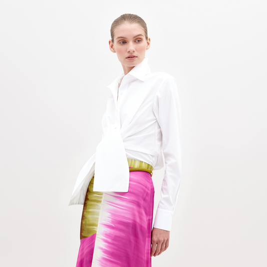 A person wearing a Mercury Blouse White and a vibrant pink skirt with green accents, standing against a plain white background, available for pre-order now. Ships by November 15th, 2024.