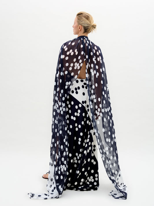 In a studio, a person models the Sabina Dress—a long black silk maxi with white polka dots and a matching cape overlay—paired with black and gold sandals.