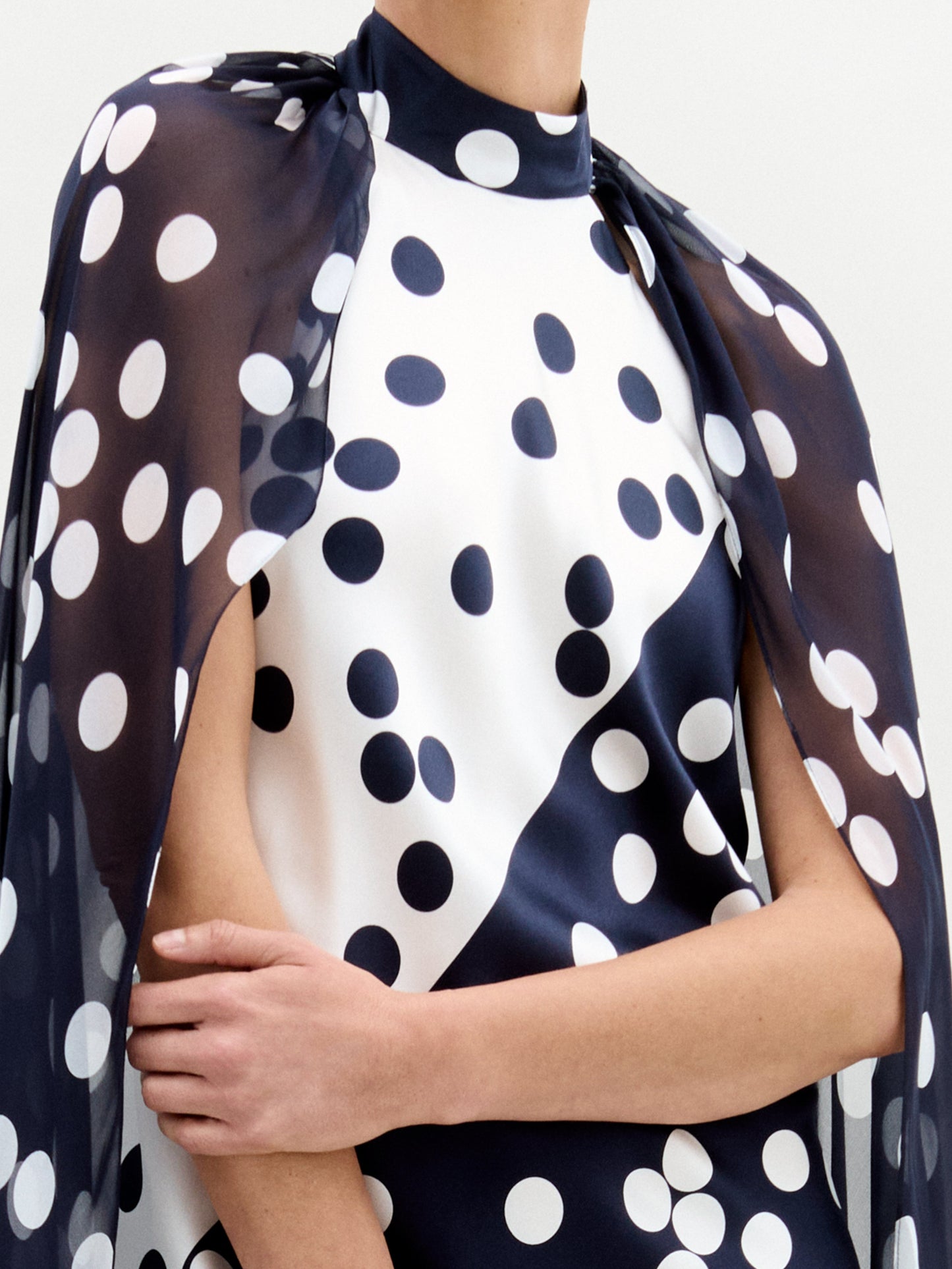In a studio, a person models the Sabina Dress—a long black silk maxi with white polka dots and a matching cape overlay—paired with black and gold sandals.