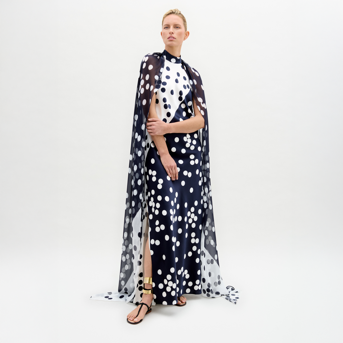 In a studio, a person models the Sabina Dress—a long black silk maxi with white polka dots and a matching cape overlay—paired with black and gold sandals.