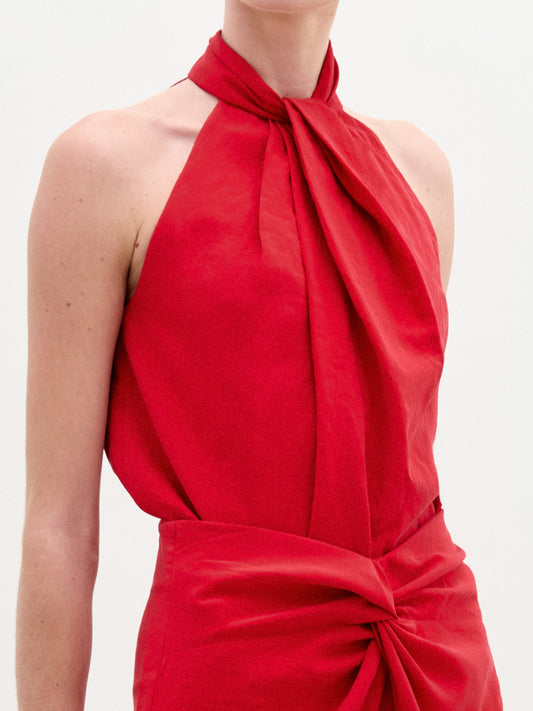 Woman wearing the Shela Blouse, a sleeveless red halter-neck top made from Italian fabric with a knotted front detail, standing against a plain light background.