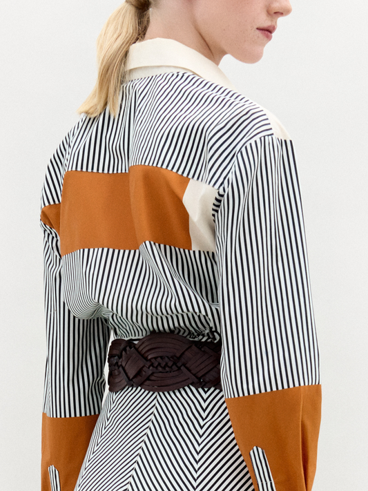 A person wearing the Soleil Blouse—long-sleeved and striped with orange and white color blocks in 100% Italian cotton—paired with a large gold belt, stands against a plain light background.