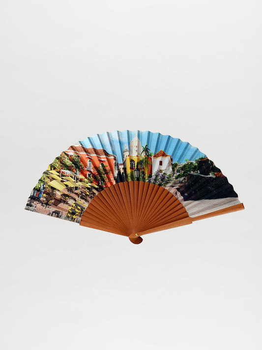 Introducing the Abanico San Juan de Dios: a delightful hand fan featuring vivid illustrations of buildings and trees on its fabric. Its frame is made from a light brown material, enhancing its sophisticated charm.
