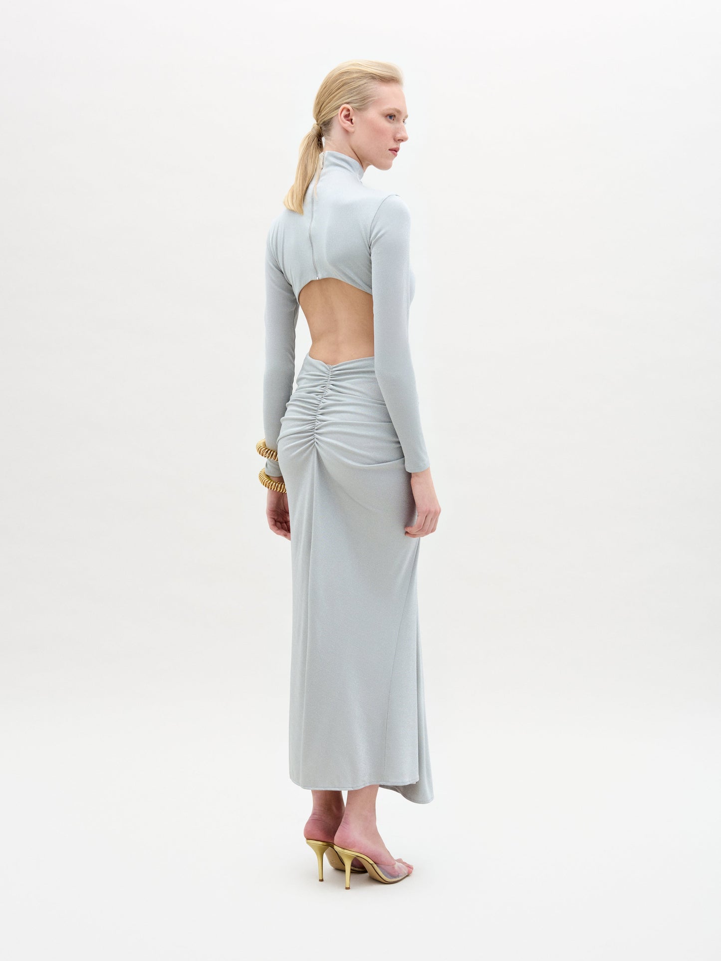 A woman in profile stands on a white background wearing the Venus Dress—a light gray, long-sleeved midi dress with an open back, ruched detailing, and a draped skirt made from Italian fabric. She pairs it with gold high heels and chunky gold bracelets.