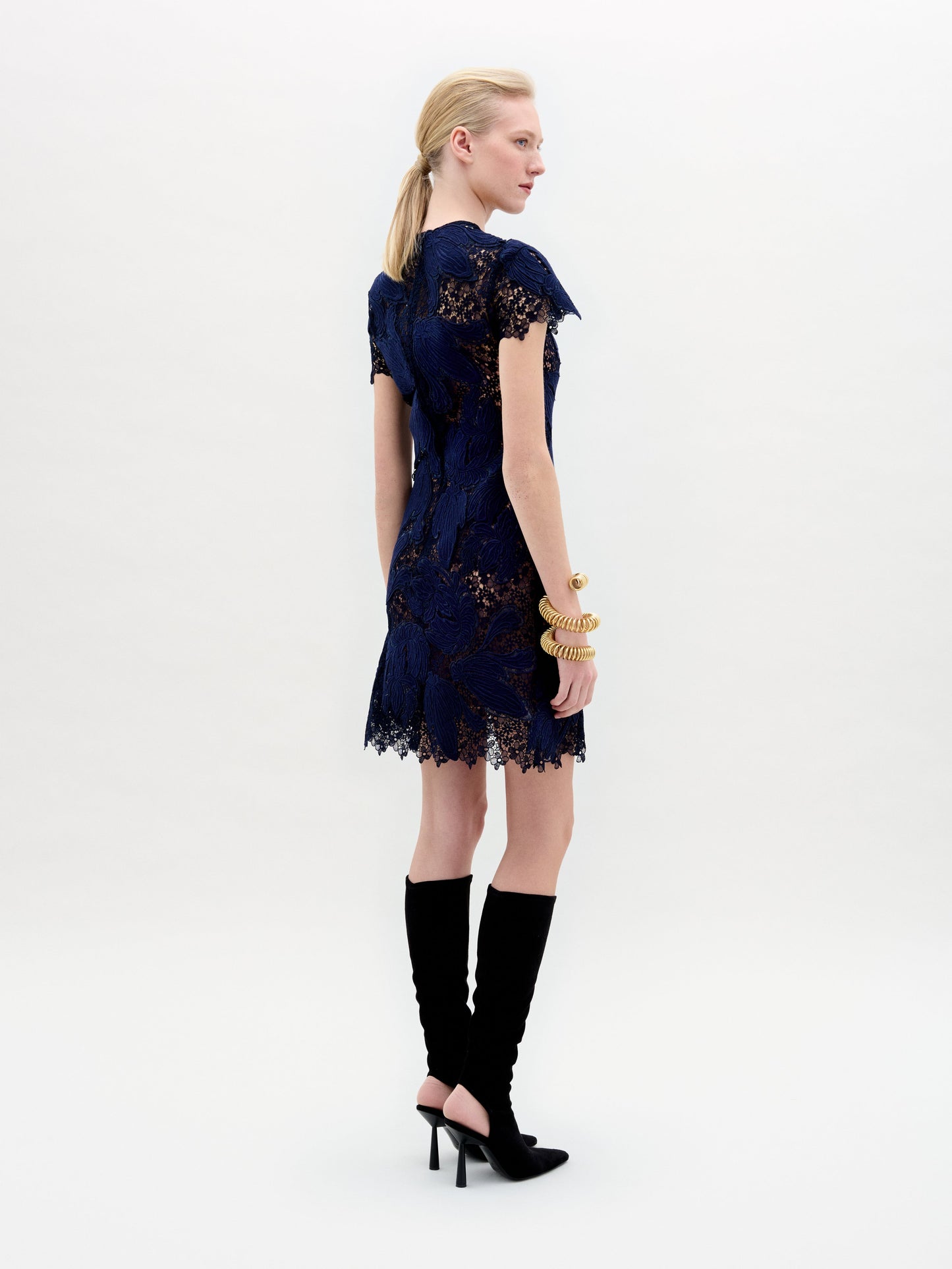 A woman stands in profile against a plain white background, wearing the Veronica Dress—a sheer lace navy blue mini dress with black detailing—paired with black knee-high heeled boots and gold bangles.