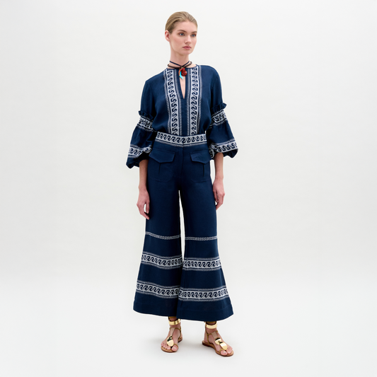 A woman stands against a plain background, wearing a navy blue embroidered blouse with ruffled sleeves and the Zina Pant—matching embroidered linen wide-leg pants—paired with gold sandals.