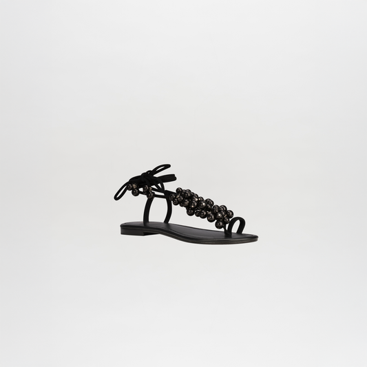 The Zaira Sandals are black leather sandals with wrap-around ankle straps and decorative beads, shown on a plain white background.