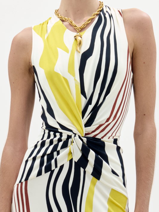 A person wearing the Luna Dress, a sleeveless midi made from Italian fabric with abstract black, yellow, and brown stripes, a twisted waist knot, criss-cross back straps, paired with a gold chain pendant necklace.