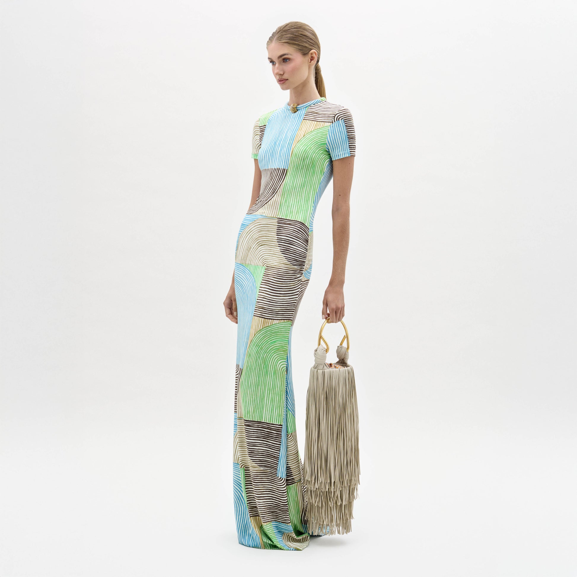 A woman in a Yara Dress, featuring multicolored patterns, stands holding a large beige fringe bag with circular handles against a plain white background.