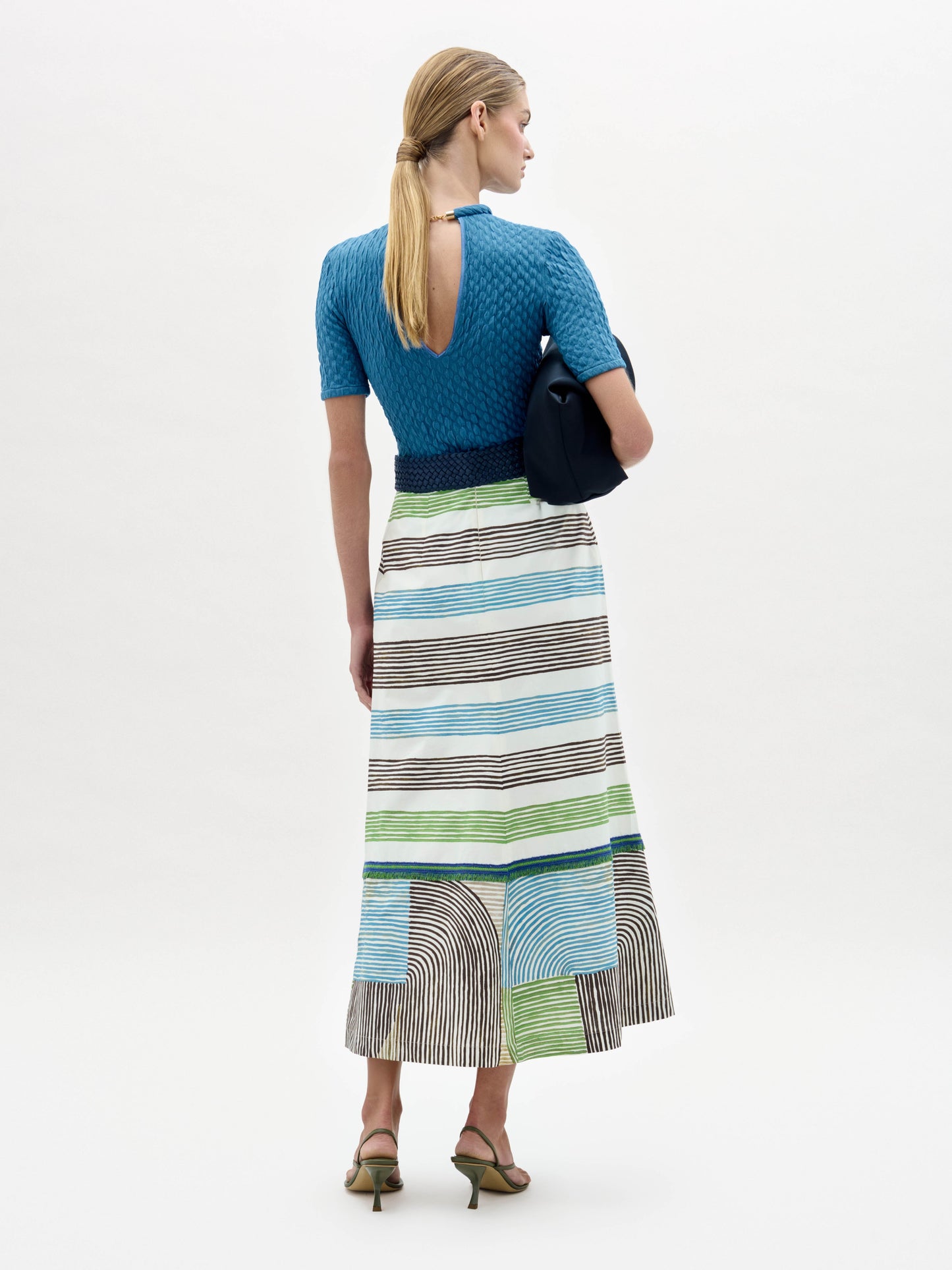 A woman faces away wearing a blue textured short-sleeve open-back top with a fitted silhouette, paired with the Lyla Skirt—a long striped midi made from Italian fabric. She carries a navy clutch and wears heeled sandals.