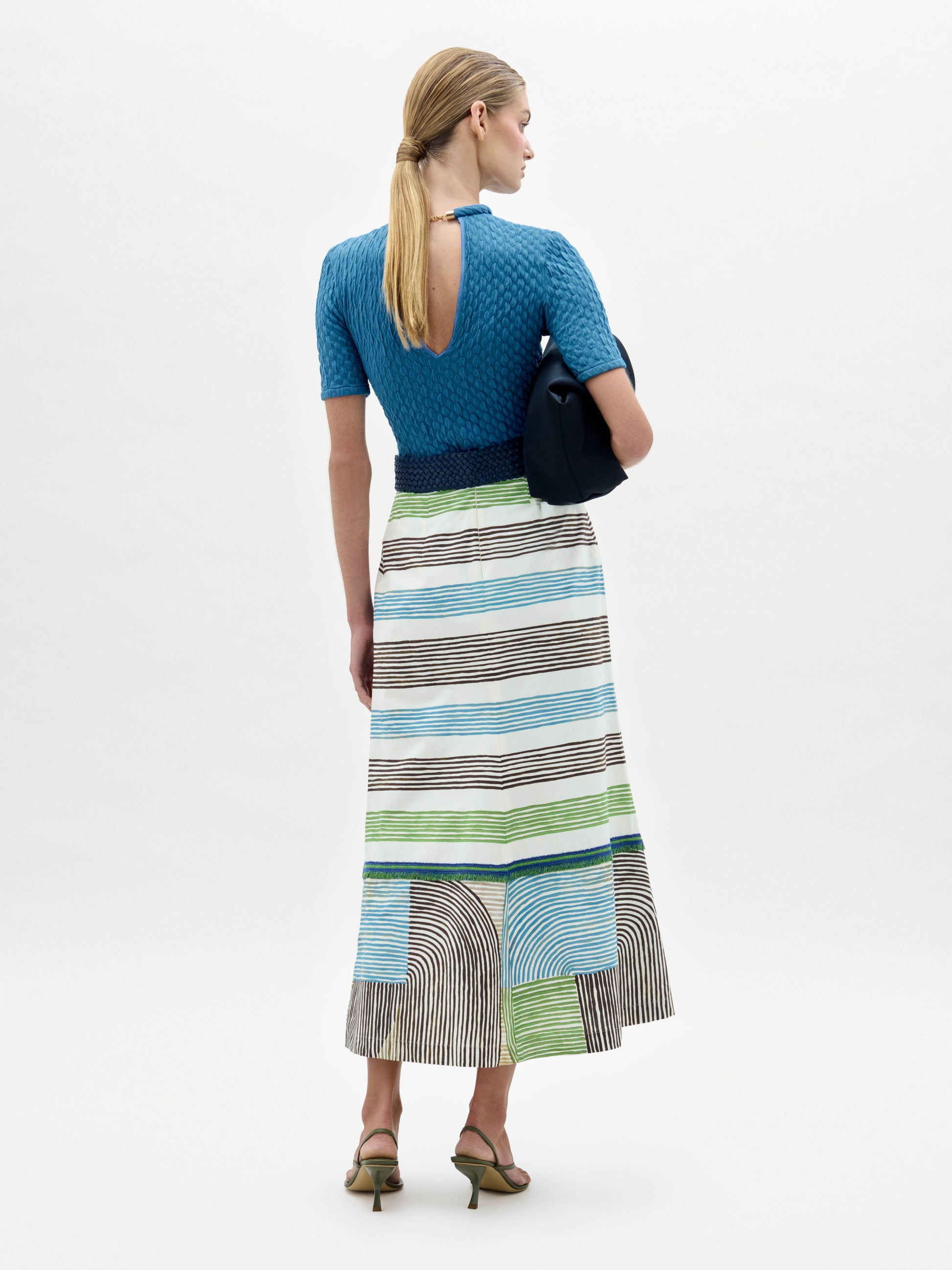 A woman faces away wearing a blue textured short-sleeve open-back top with a fitted silhouette, paired with the Lyla Skirt—a long striped midi made from Italian fabric. She carries a navy clutch and wears heeled sandals.