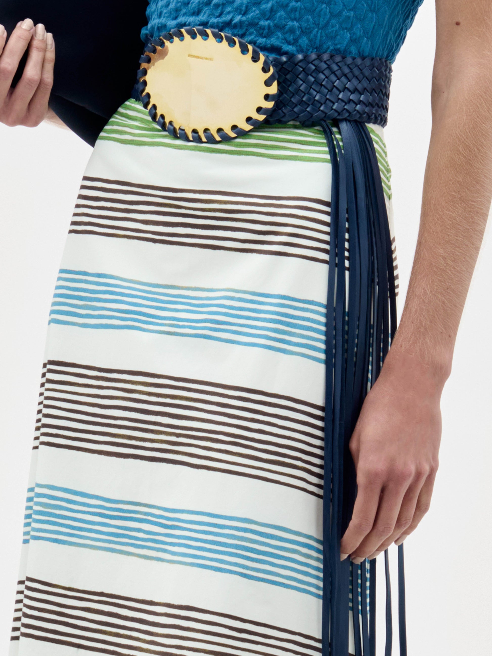 A person wears the Lyla Skirt, featuring white fabric with blue, green, and brown horizontal stripes, paired with a blue textured top and a wide dark blue woven belt with a large oval buckle. Their right hand rests at their side.