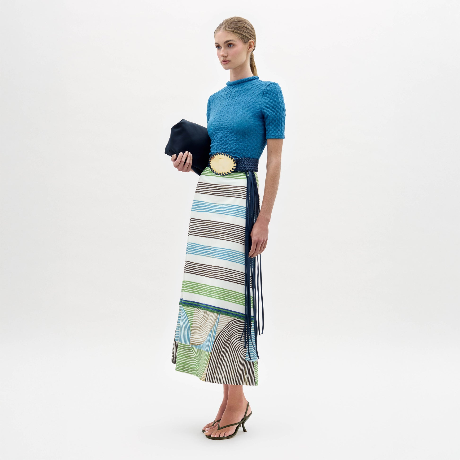 A woman stands against a white background wearing a blue short-sleeve top, the Lyla Skirt—featuring a fitted striped midi design in blue, green, and brown—a wide black belt with gold buckle, black sandals, and holding a black clutch.
