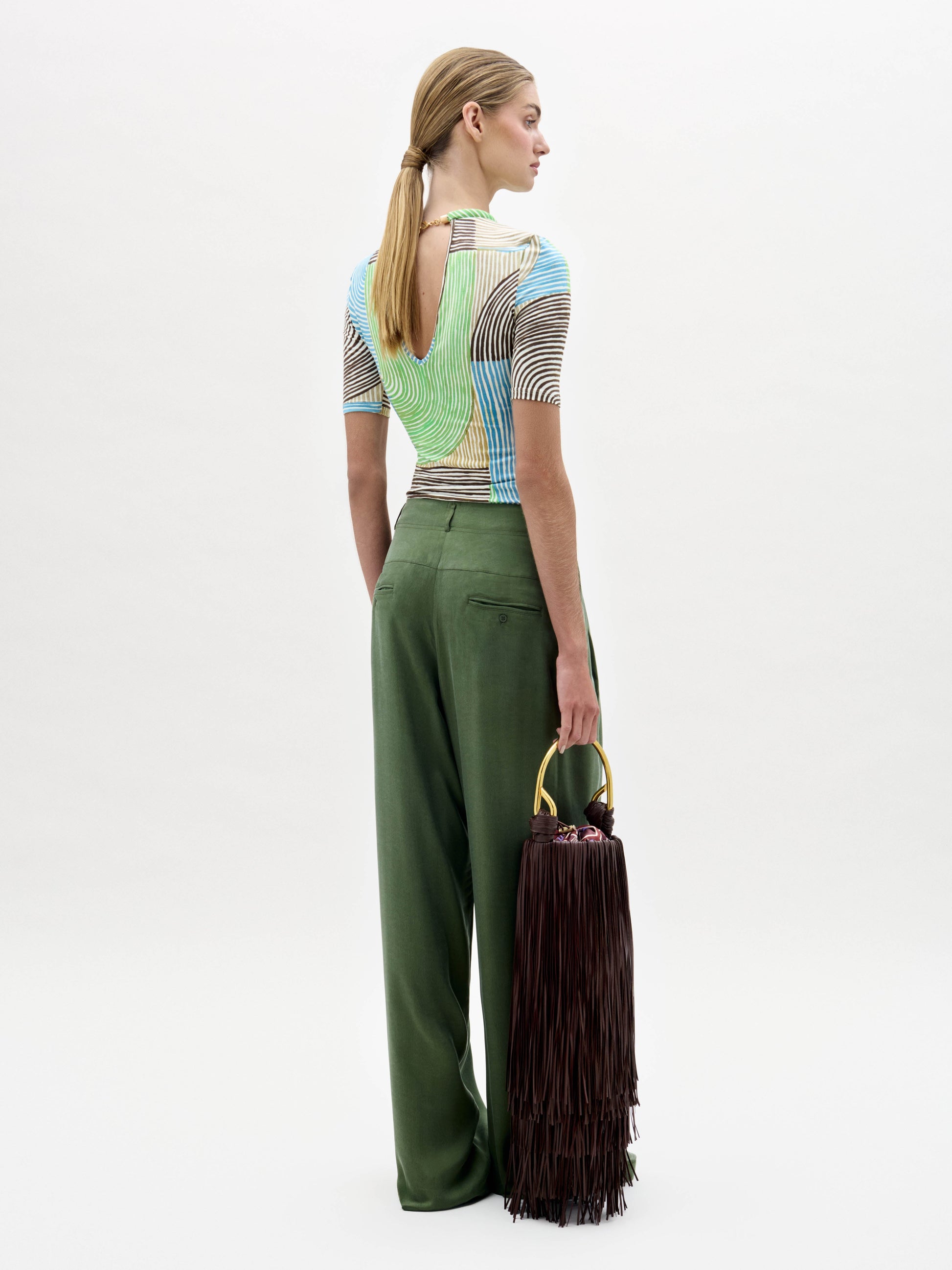 A woman stands against a white background wearing wide-leg green Opal Pants, a colorful striped top, and holding a large dark fringed bag.